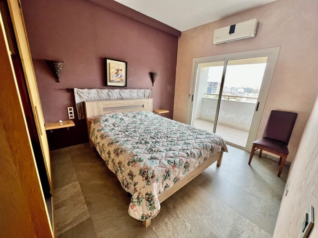 Une chambre à coucher comprenant un lit, une chaise, une table de chevet, un tableau au mur et une porte coulissante donnant sur un balcon.