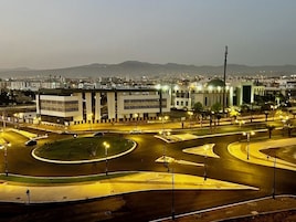 Ein gut beleuchteter Kreisverkehr mit mehreren Fahrbahnen und einer Mittelinsel, umgeben von Gebäuden und einem fernen Bergpanorama.