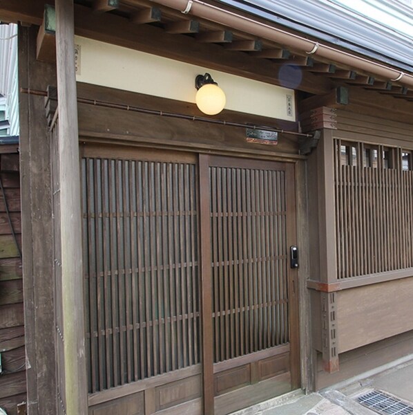A traditional Japanese wooden gate with a sliding door, a small light fixture above it, and a metal fence to the right.
