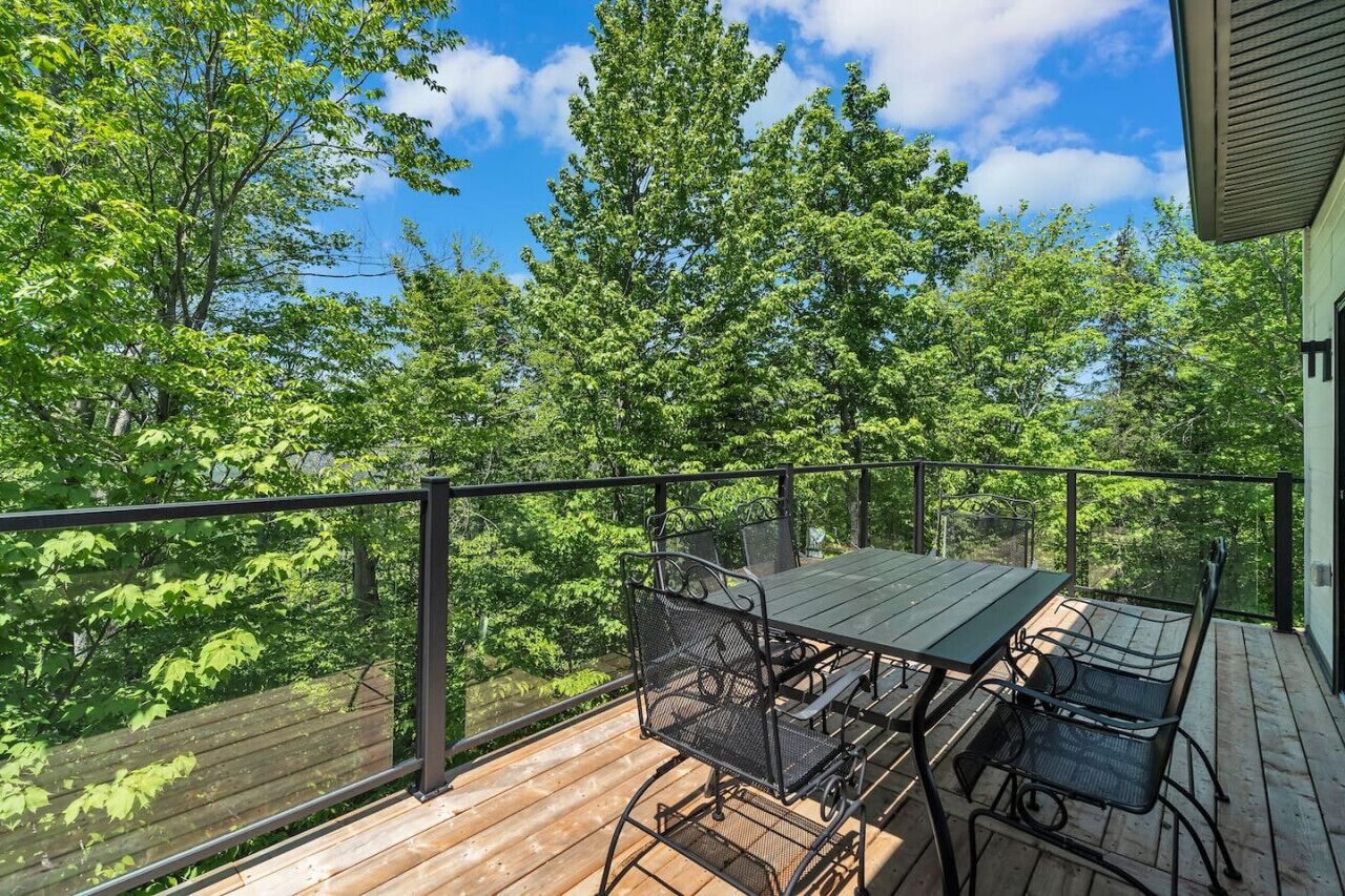 A wooden deck with a table and chairs, surrounded by a metal railing, overlooking a forested area.