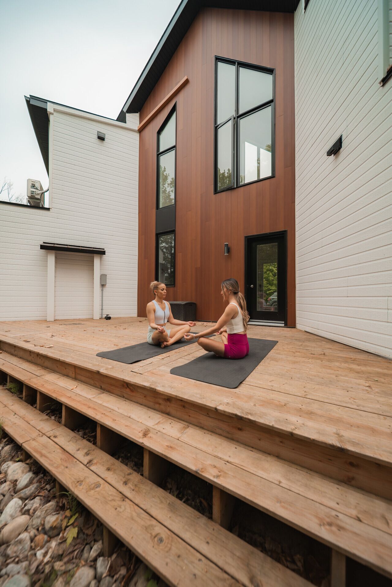 A modern house with a wooden deck and two people sitting on yoga mats.
