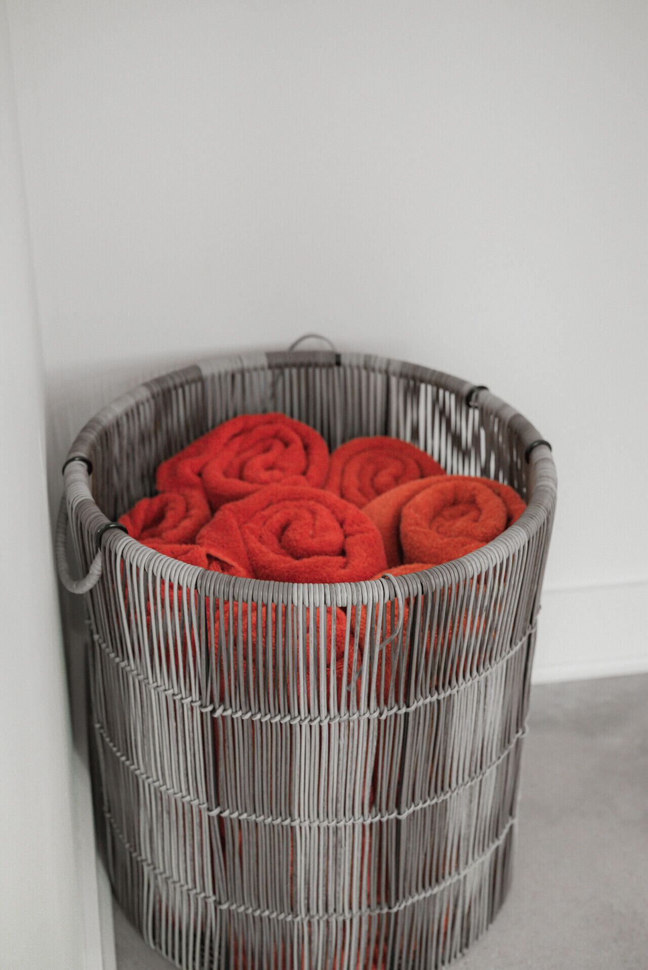 Un panier à linge en osier gris, rempli de serviettes rouges soigneusement pliées, est posé contre un mur blanc.