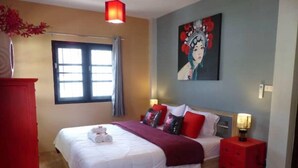 A bedroom with a bed, a nightstand, a red dresser, a window with curtains, and a painting on the wall.