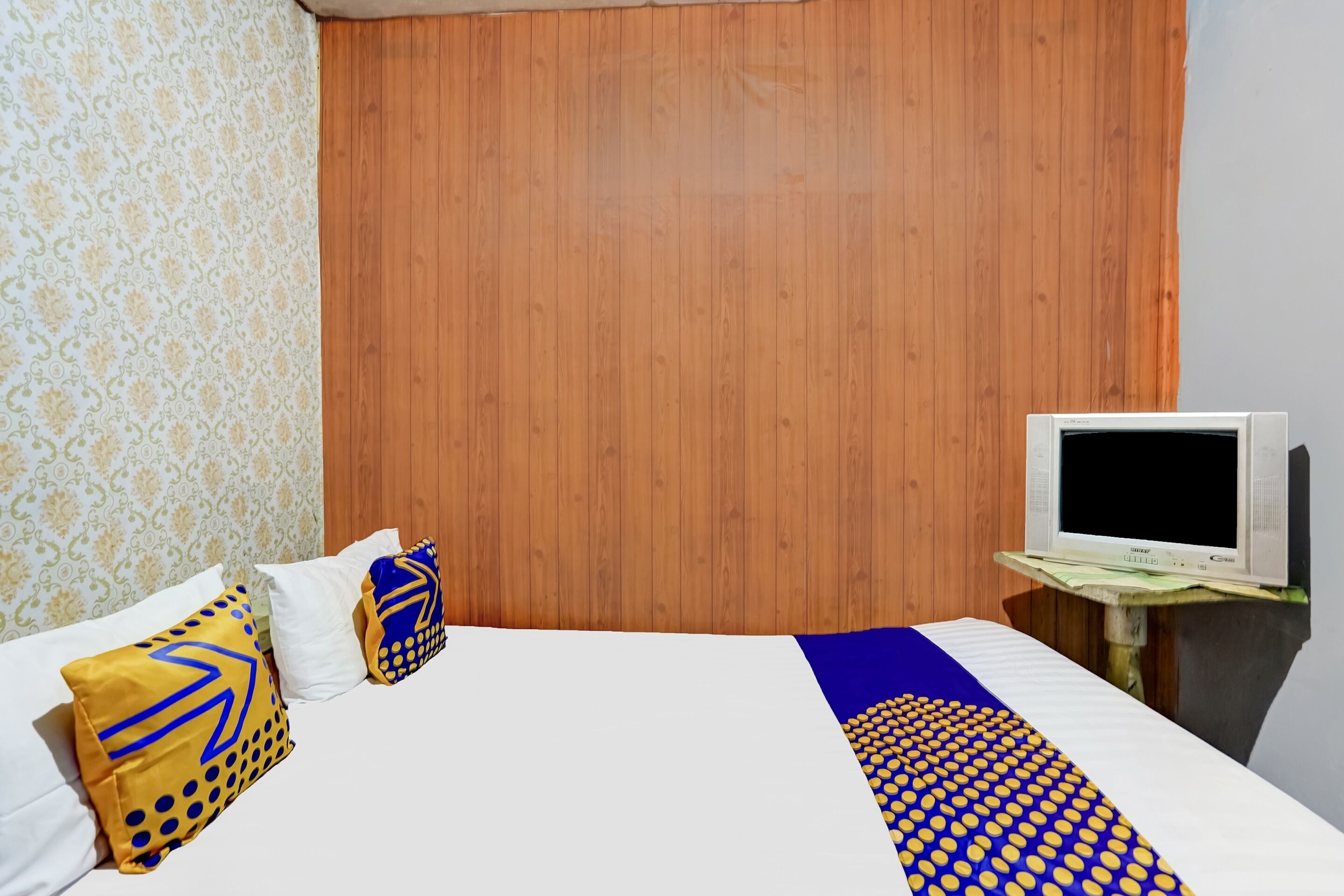 A hotel room with a wooden headboard, a bed with patterned pillows, a TV on a wooden stand, and a patterned wall.