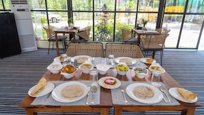 Una mesa de comedor dispuesta con diversos platos y cubiertos, rodeada de sillas de mimbre, en una habitación con ventanas amplias y una alfombra a rayas.