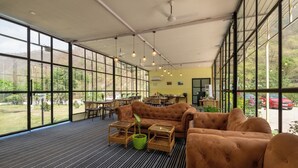 Un espacio interior amplio con ventanas grandes, un sofá marrón, mesas de mimbre y una planta verde en maceta.