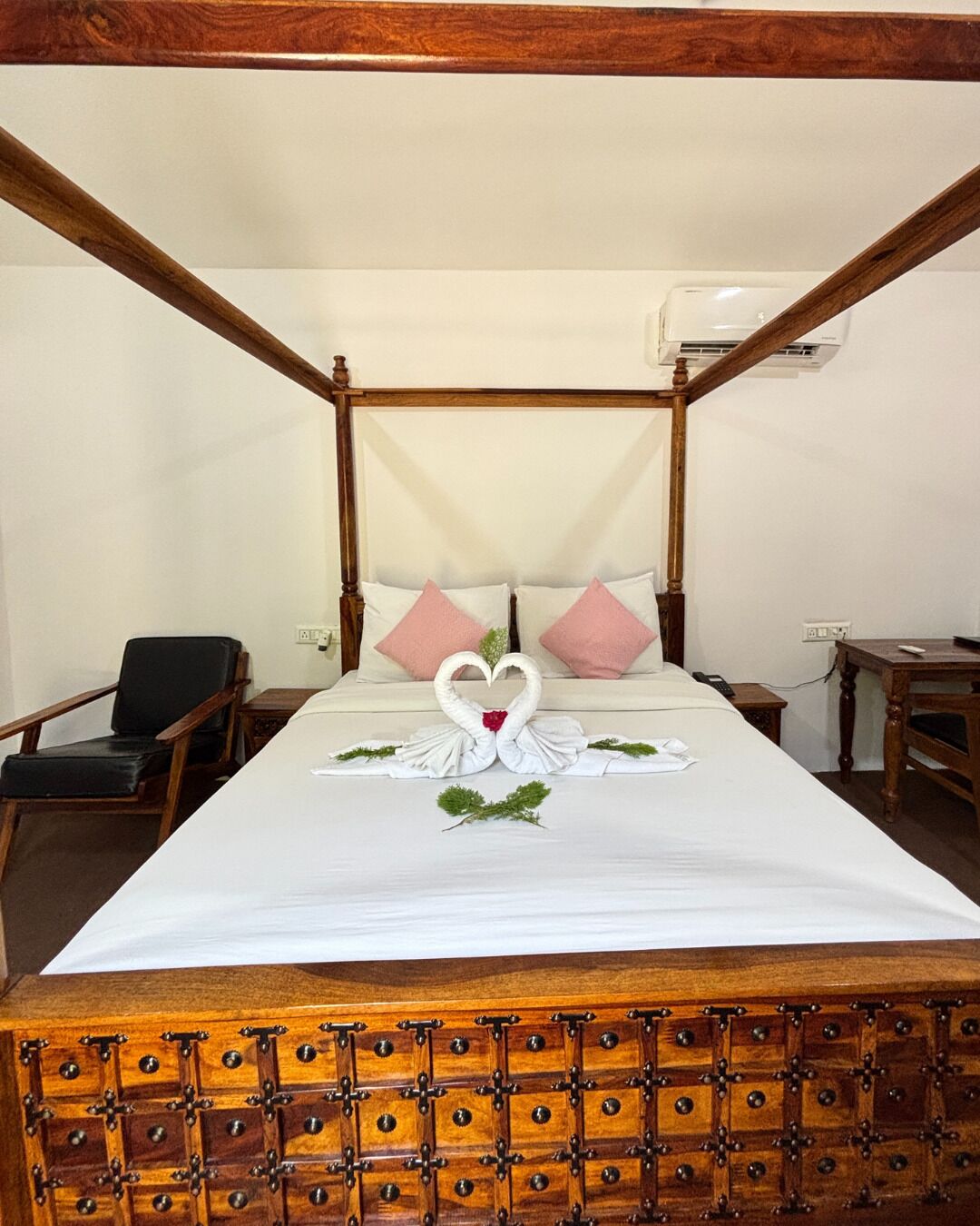 A four-poster bed with a white bedspread and a swan decoration. There are two pillows, one pink and one white. A chair and a small table are visible in the room.
