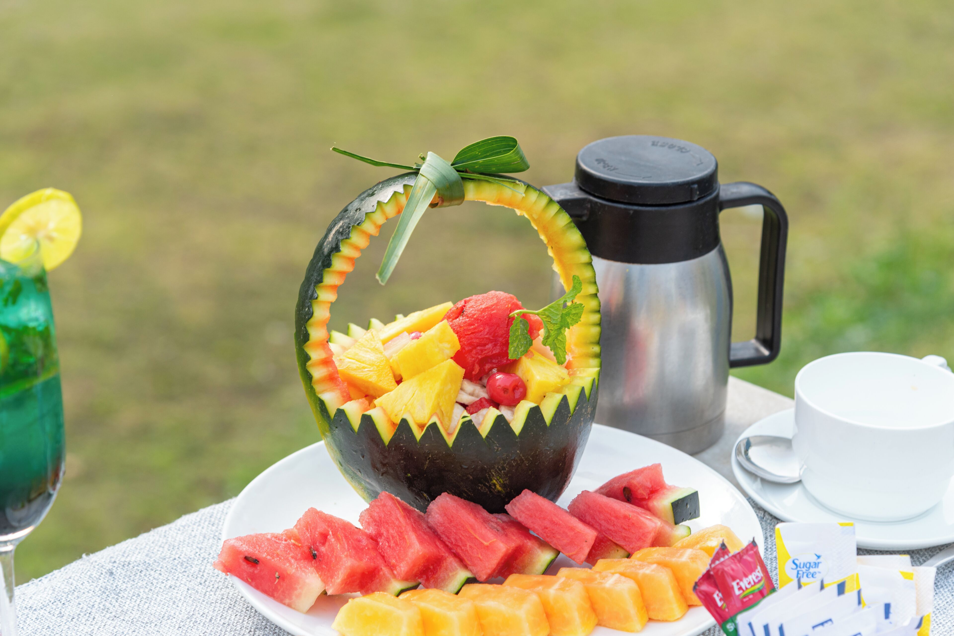 A fruit salad in a hollowed-out watermelon bowl, a metal coffee pot, a white cup, and sliced watermelon on a table outdoors.