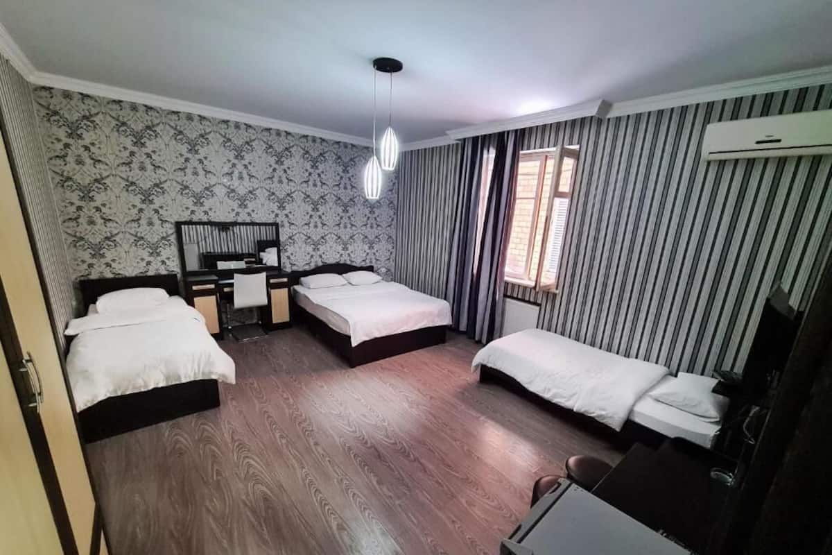 Ein Hotelzimmer mit drei Betten, einem Schreibtisch und einem Stuhl. Ein Fenster mit Vorhängen und eine Klimaanlage befinden sich an der Wand.