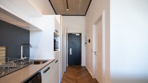 A narrow hallway with a wooden floor, a kitchen area with a dishwasher and a sink, and a door leading to another room.