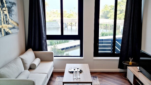 A modern hotel room with a bed, a sofa, a coffee table, and a TV. There are two windows with curtains, and a painting on the wall.