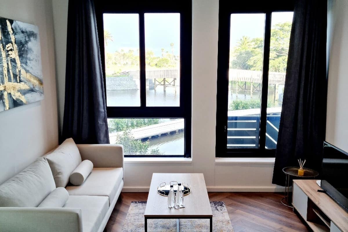 A modern hotel room with a bed, a sofa, a coffee table, and a TV. There are two windows with curtains, and a painting on the wall.