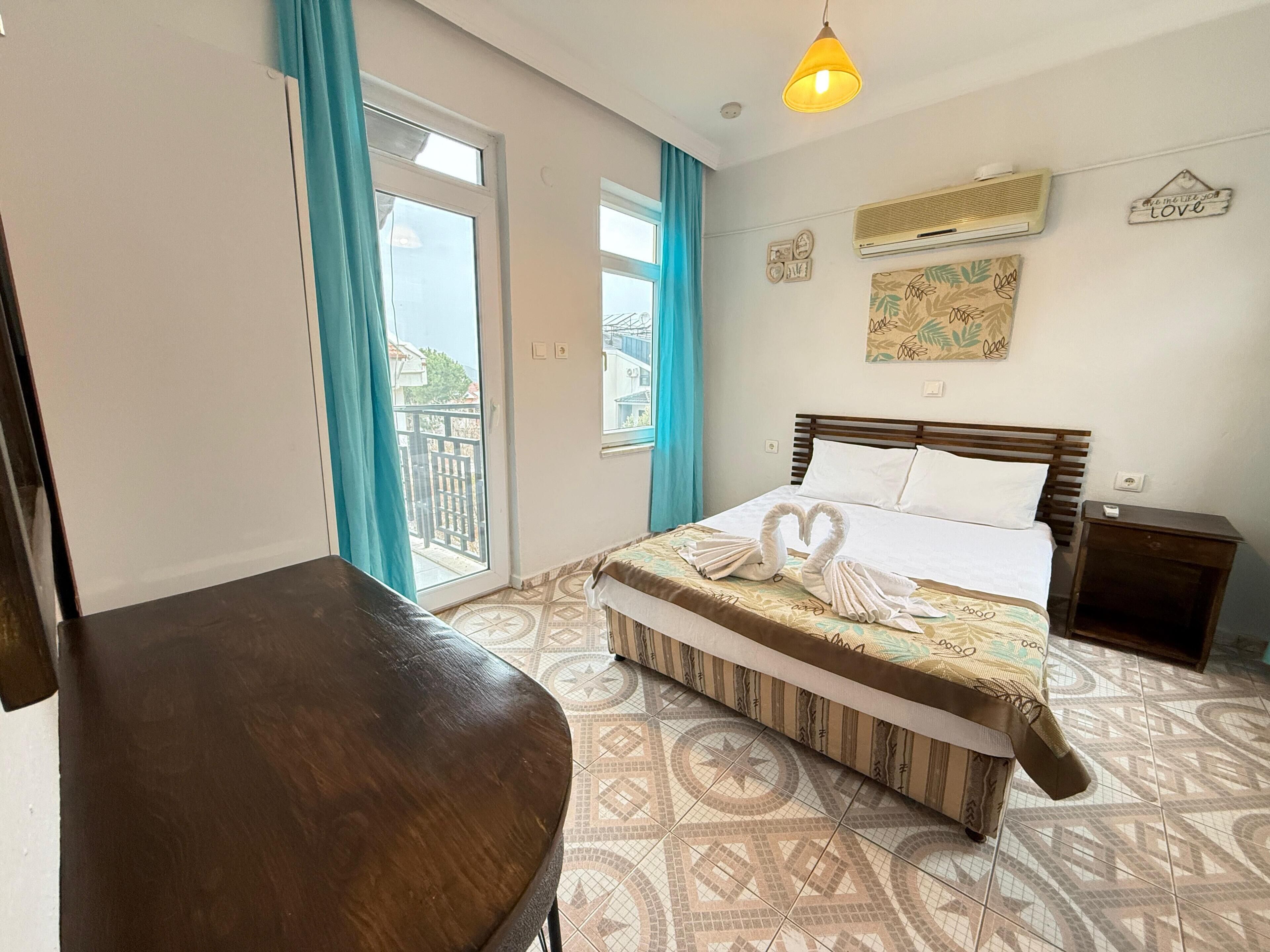 Habitación de hotel con una cama, una silla de madera, un balcón con cortinas, un aire acondicionado instalado en la pared y un letrero decorativo.