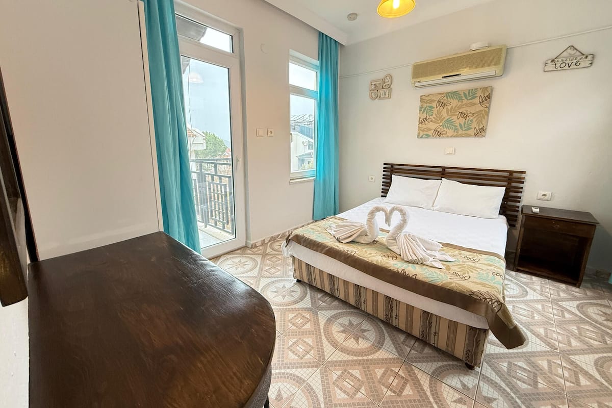 A hotel room with a bed, a wooden chair, a balcony with curtains, a wall-mounted air conditioner, and a decorative sign.