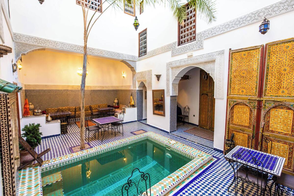 Una piscina interior con suelo de baldosas, un área de descanso con mesas y sillas, y puertas decorativas.