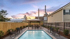 A rectangular swimming pool with a wooden deck and chairs, surrounded by a wooden fence, with a sunset and industrial buildings in the background. - Remarkable Superdome Group Home w/Heated Pool, Parking, Patio, 8 Beds & 5 Baths (New Orleans)