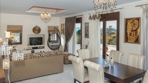 A modern living room with a sofa, dining table, and chairs. There is a television, a chandelier, and artwork on the walls. - Heliopetra Lux Villa with private Pool (Chania)