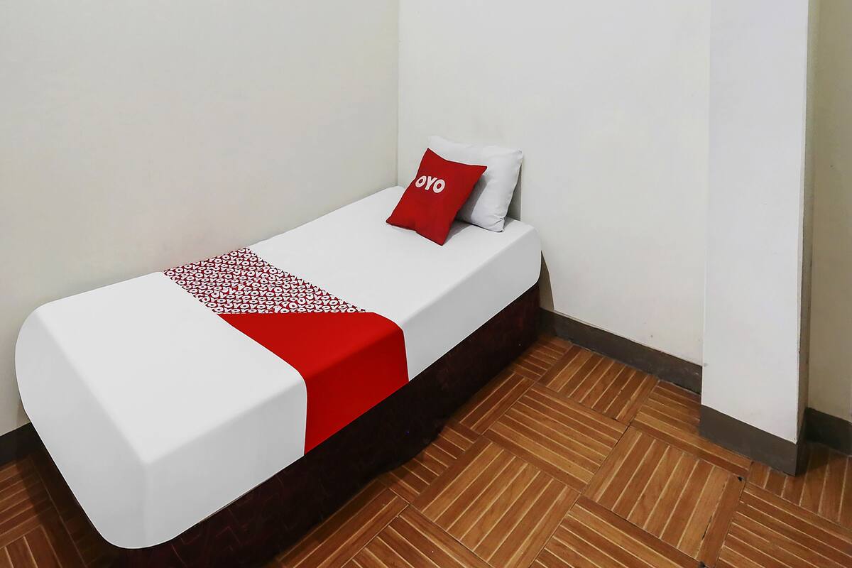 Una cama individual con colchón blanco y manta roja, un almohadón rojo con la palabra "OYO" y suelo de madera.