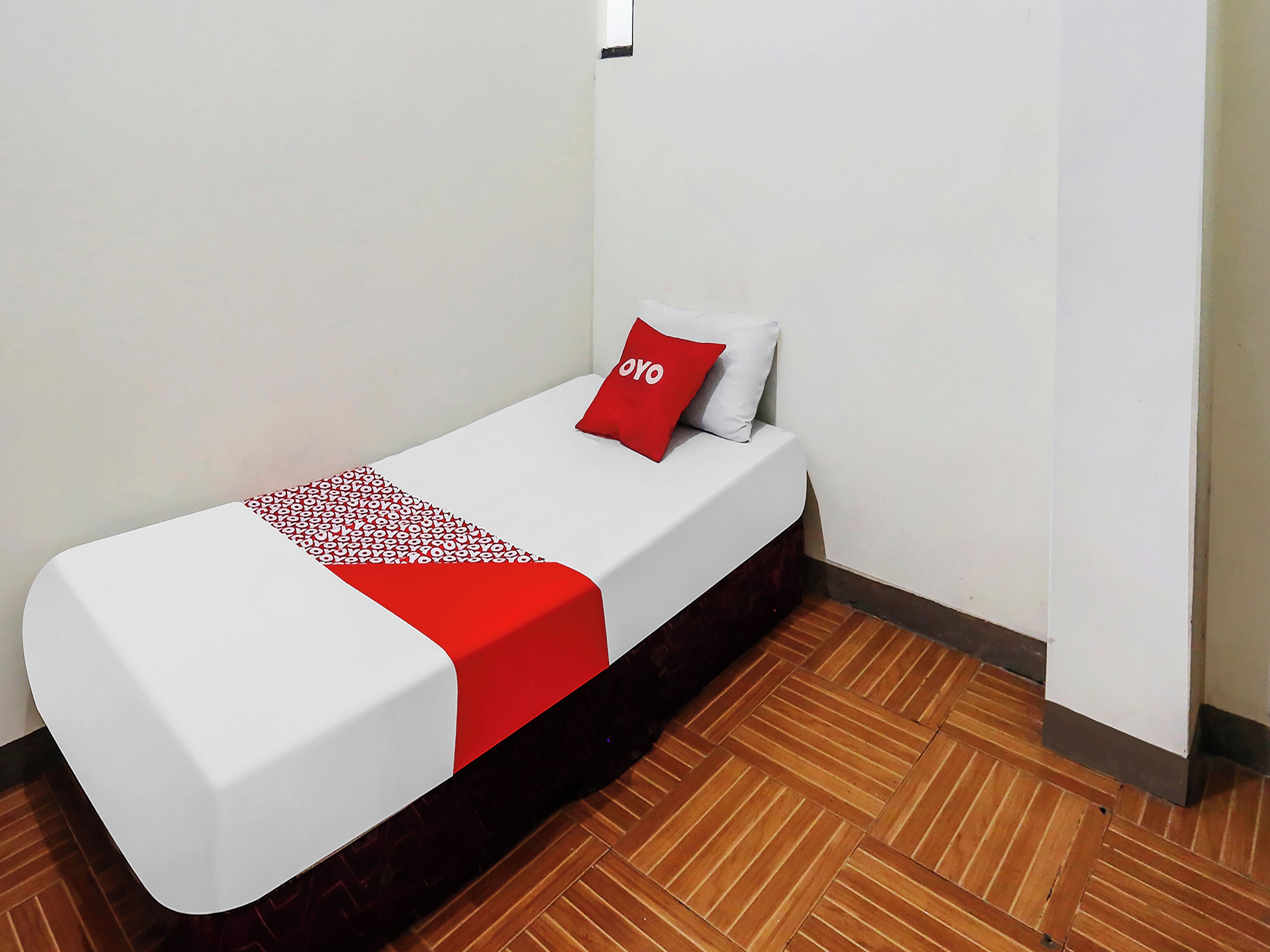 A single bed with a white mattress and a red blanket, a red pillow with the word "OYO" on it, and a wooden floor.