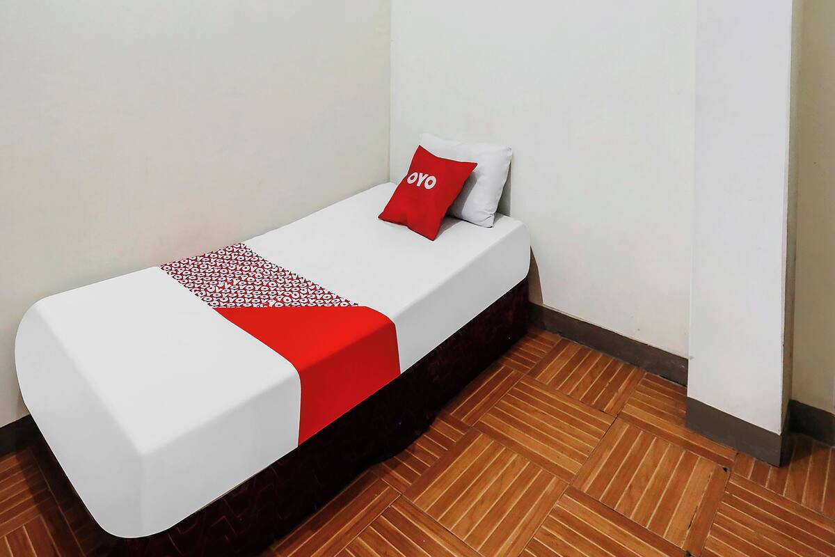 Una cama individual con colchón blanco y manta roja, un almohadón rojo con la palabra "OYO" y suelo de madera.