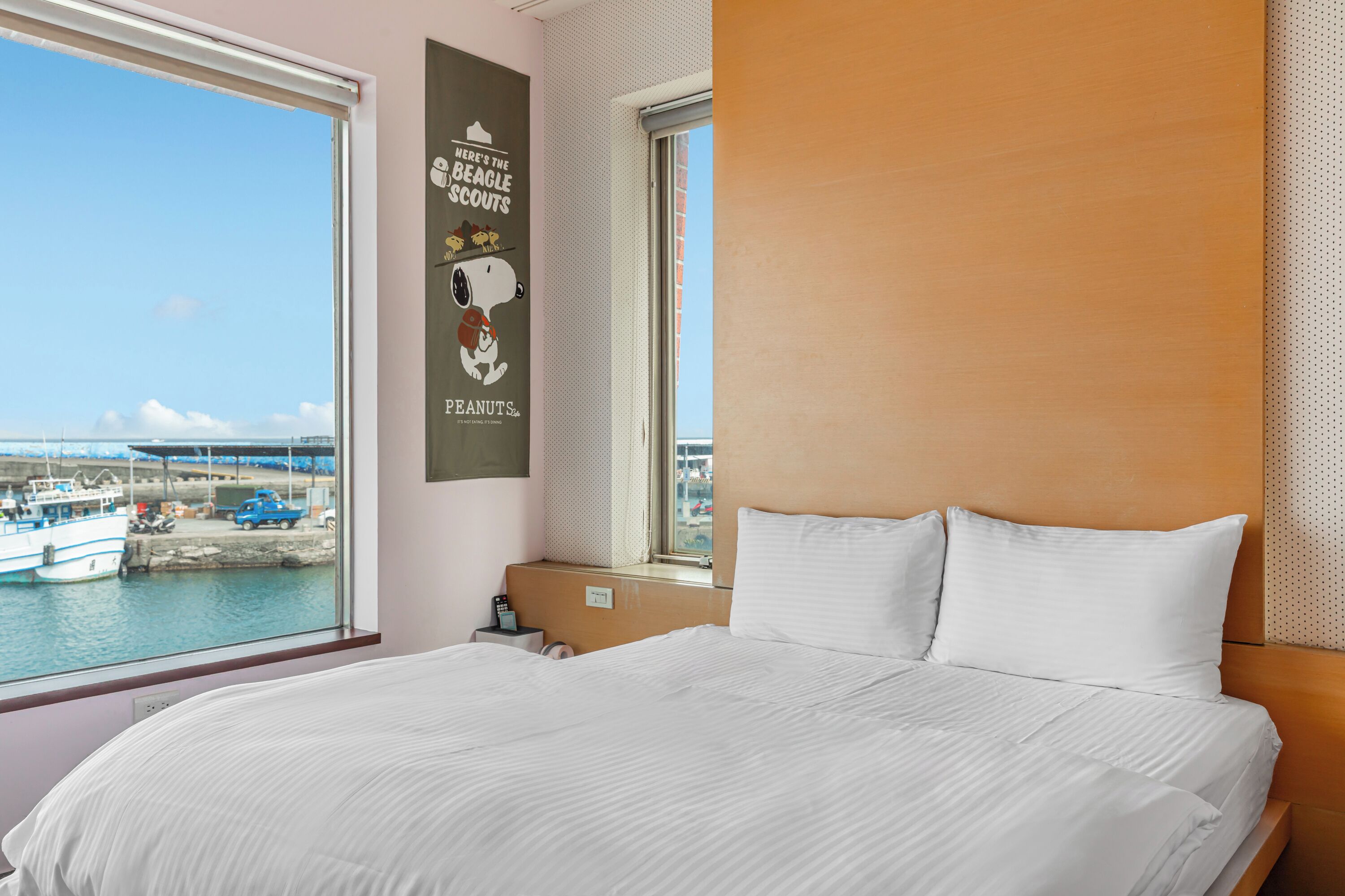 A hotel room with a large bed, a window with a view of boats and a pier, and a wall poster with a dog and text.