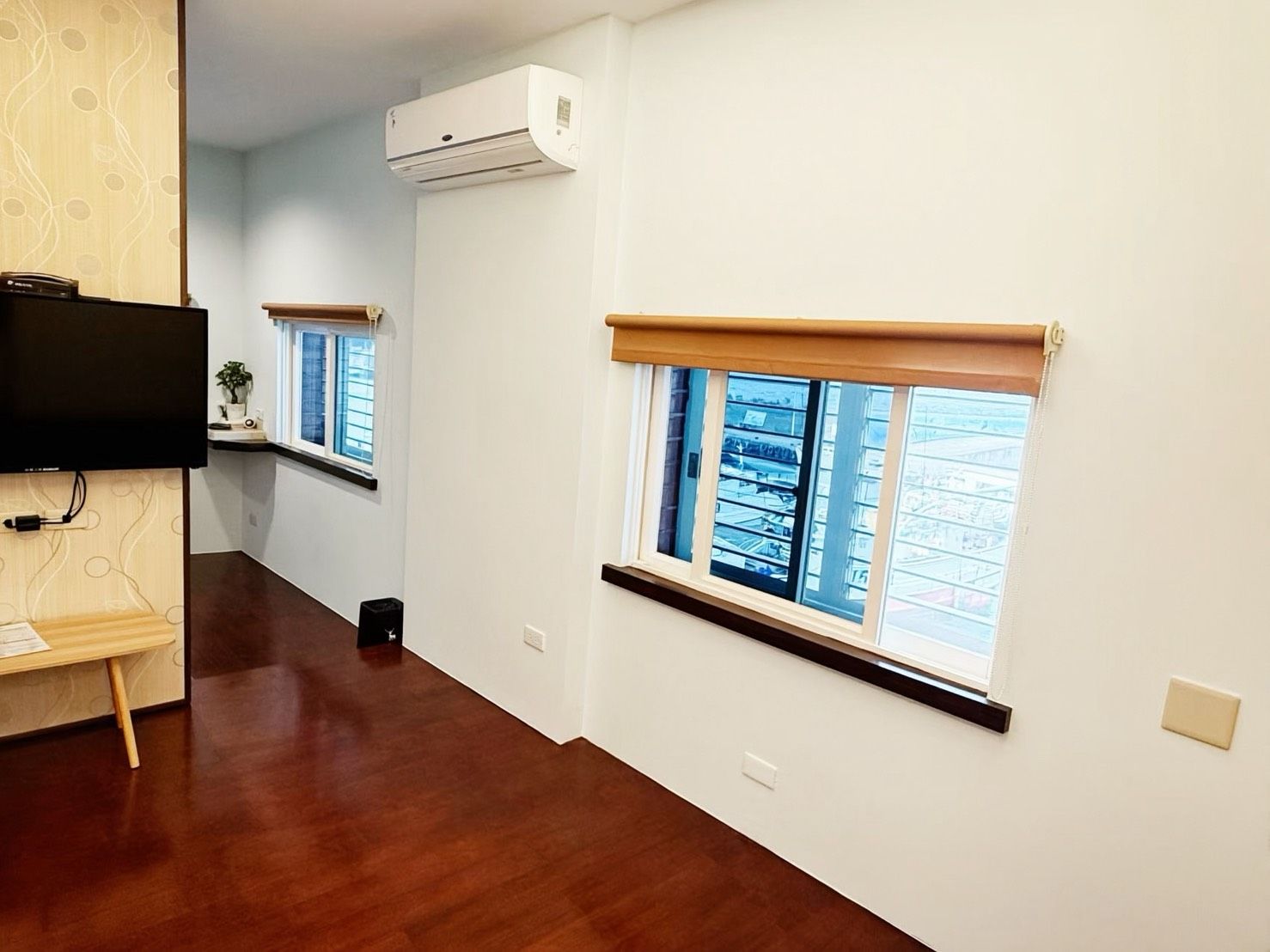 A hallway with wooden flooring, white walls, and a mounted air conditioner. There are windows with blinds and a television on a stand.
