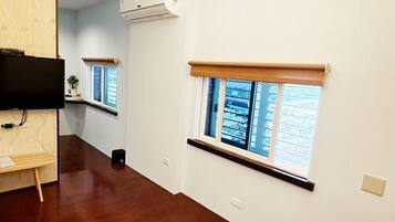 A hallway with wooden flooring, white walls, and a mounted air conditioner. There are windows with blinds and a television on a stand.