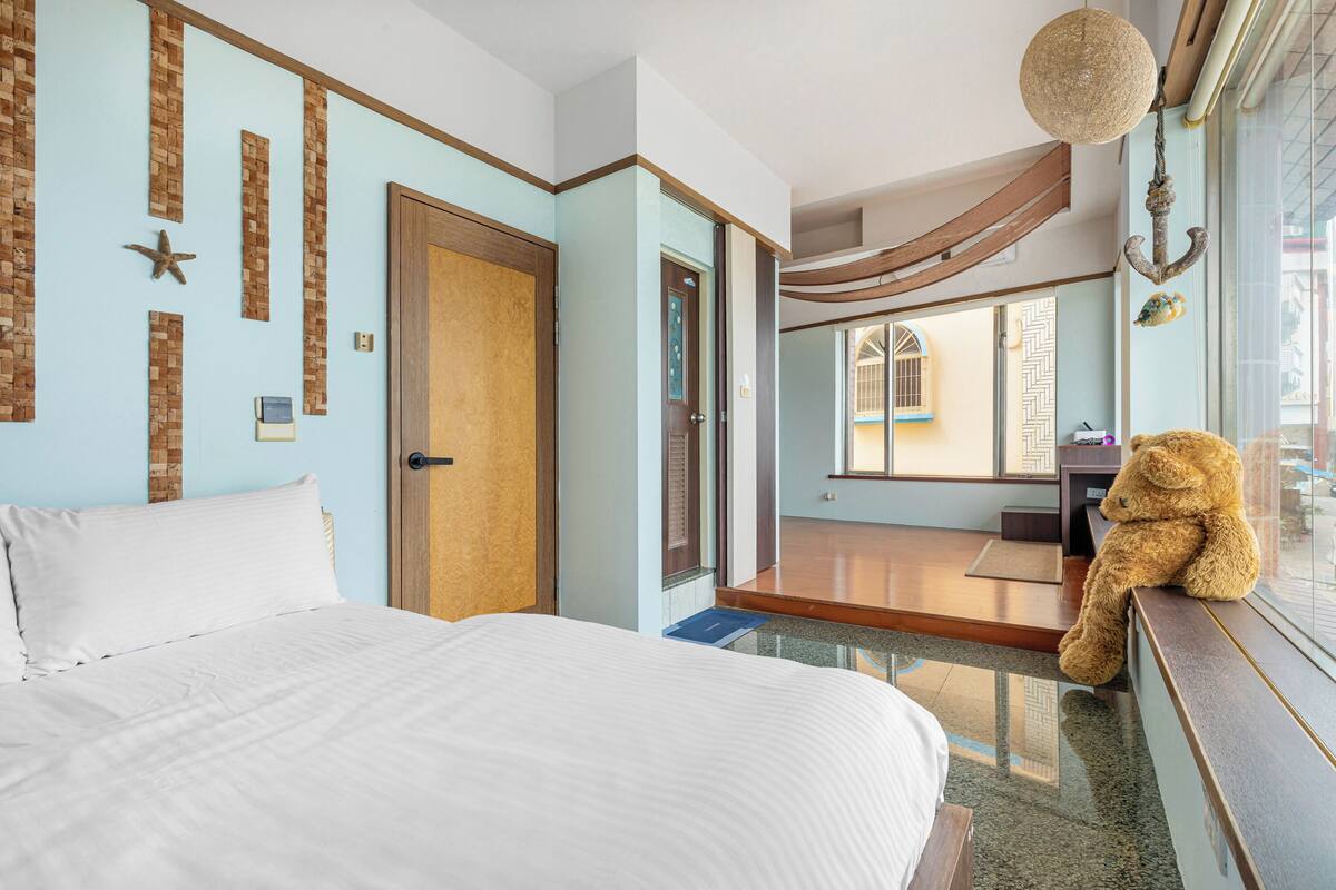 A hotel room with a bed, a teddy bear on the window sill, and a view of the city through the window.