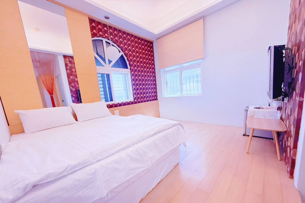 A bedroom with a bed, a bedside table, a mirror, and a window.