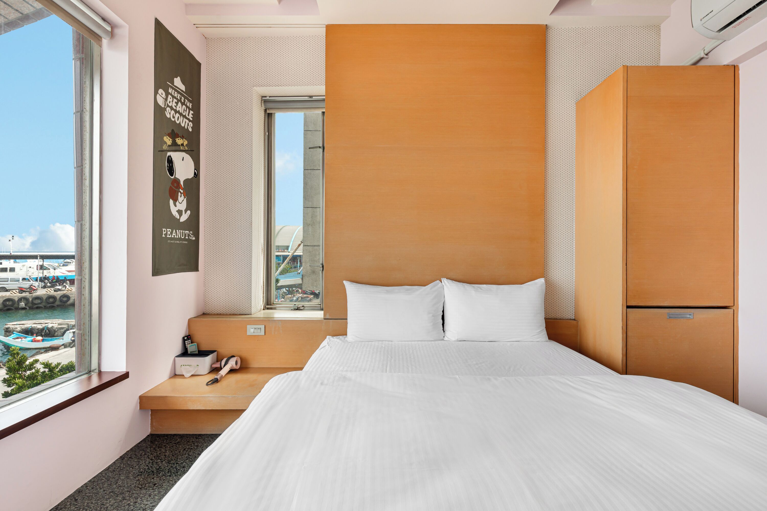 A hotel room with a large bed, a wooden wardrobe, a window with a view of boats, and a poster on the wall.