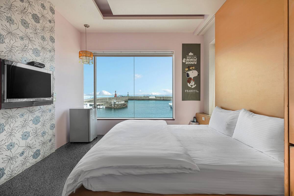 A hotel room with a large bed, a TV mounted on the wall, a small fridge, and a window with a view of the sea and a lighthouse.