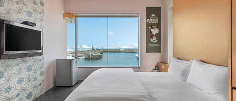 Ein Hotelzimmer mit einem großen Bett, einem an der Wand befestigten Fernseher, einer kleinen Minibar und einem Fenster mit Blick auf das Meer und einen Leuchtturm.