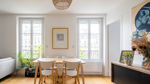 Uma sala de jantar com uma mesa e cadeiras de madeira, um sofá branco e uma TV sobre um suporte preto.