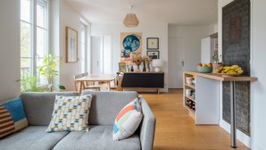 Uma sala de estar moderna com um sofá cinzento, uma mesa de jantar de madeira e uma cozinha com uma fruteira e objetos decorativos.