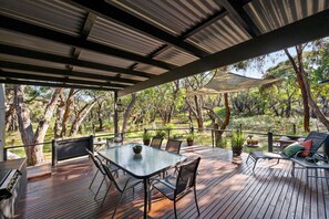 Una terraza exterior con mesa y sillas, rodeada de árboles y una zona cubierta con techo metálico.