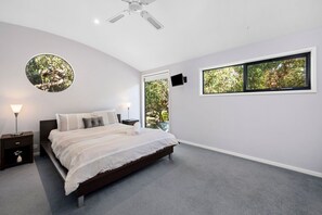 Un dormitorio con cama, mesita de noche, lámpara, ventilador de techo y ventana con vista a árboles.