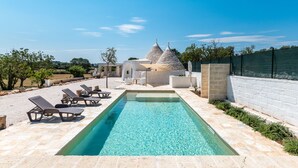 A swimming pool with lounge chairs, a stone building with conical roofs, and a clear blue sky. - Trullo La Dolina by Wonderful Italy (Ostuni)