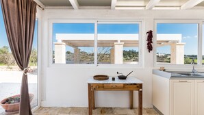 A kitchen with a white countertop, a wooden table, and a window with a view of the landscape. - Trullo La Dolina by Wonderful Italy (Ostuni)