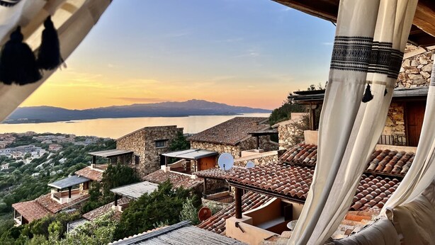 A view of a coastal town with stone buildings and terracotta roofs, overlooking a body of water and distant mountains. - AQUA GREEN HOUSE (Porto Rotondo)