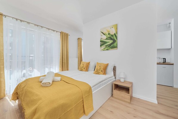 A modern bedroom with a bed, a bedside table, a nightstand, a painting, and a view of a kitchen area.