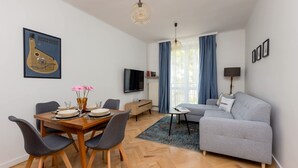 A modern living room with a grey sofa, a dining table with chairs, a TV mounted on the wall, and a large window with blue curtains. - Comfortable Apartment in Downtown | A place to work (Warszawa)