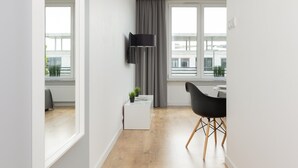 Habitación moderna y bien iluminada con un amplio ventanal, un televisor de pantalla plana en la pared, una pequeña mesita blanca con una planta y una silla negra con patas de madera.