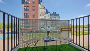 A balcony with artificial grass, two wooden benches, a small table with a vase of flowers, and a view of a multi-story building. - Spacious Apartment | Balcony, Parking, Desk (Wroclaw)