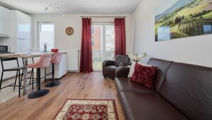 A modern living room with a brown leather sofa, a dining area with a table and chairs, a kitchen with white cabinets, and a large window with red curtains. - Spacious Apartment | Balcony, Parking, Desk (Wroclaw)