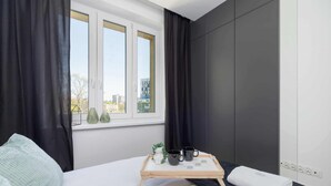 Una habitación de hotel moderna con una cama, un ventanal con cortinas, una mesita de noche con una bandeja que contiene tazas y una pequeña planta, y vistas a la ciudad.