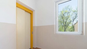Una habitación con una puerta blanca de marco amarillo, una ventana que da a árboles y un piso de baldosas.