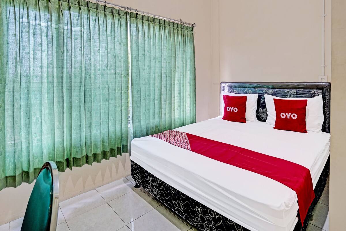 Un letto rifatto con cura, coperto da una trapunta bianca e rossa, due cuscini rossi con la scritta "OYO" e una tenda verde.