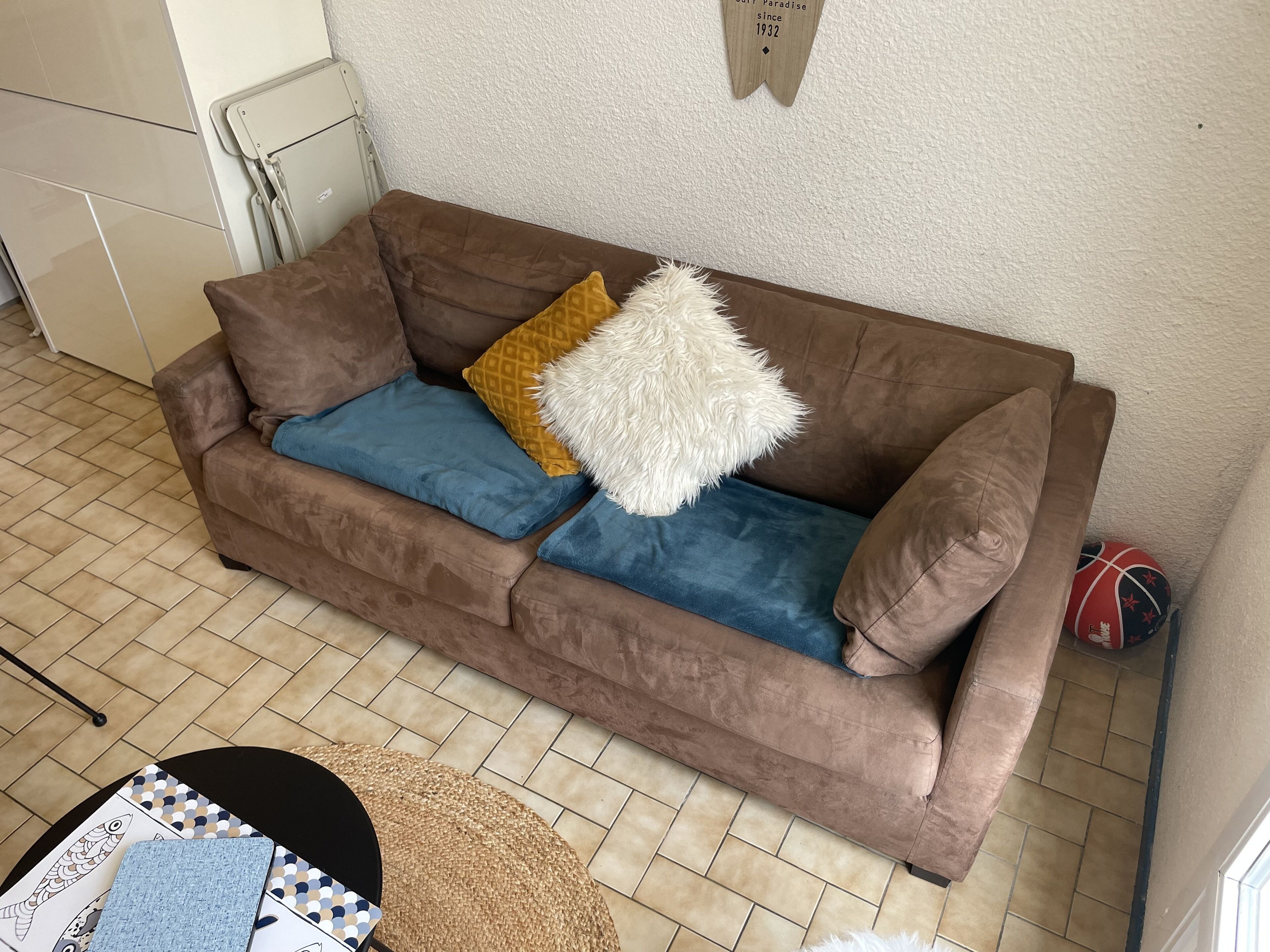 Un canapé marron avec des coussins bleus et jaunes, un oreiller blanc et moelleux, et un petit ballon de basket dans le coin.