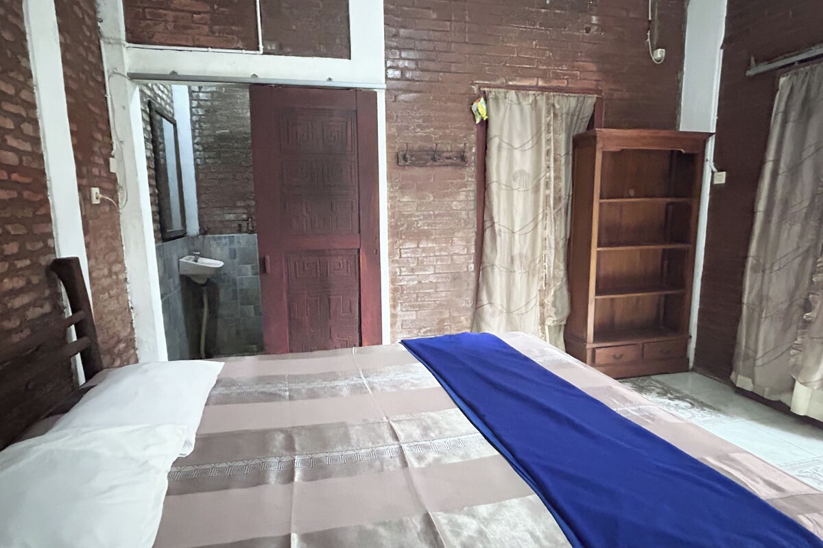 A room with a bed, a wooden wardrobe, a bathroom with a sink, and a window with curtains.