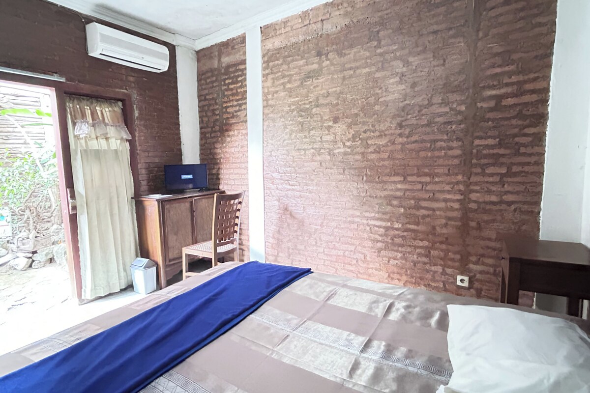 A room with a bed, a desk, and a computer. There is a brick wall and a window with curtains.